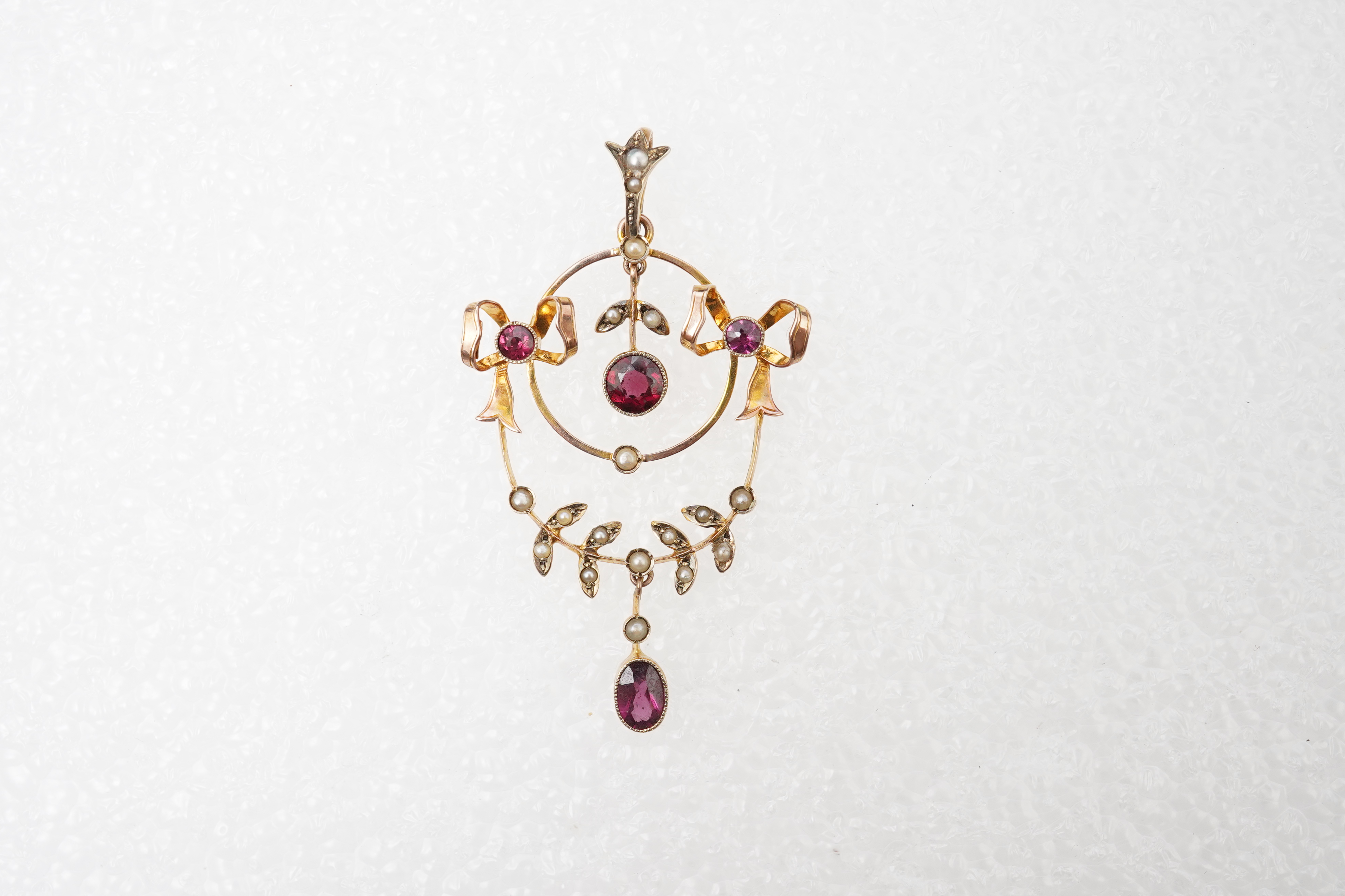 An Edwardian garnet and seed pearl pendant, early 20th century, of foliate design with ribbon bow motifs, set with circular-cut and oval garnets and seed pearls, mounted in gold, length 5.2cm, stamped 9CT, gross weight a